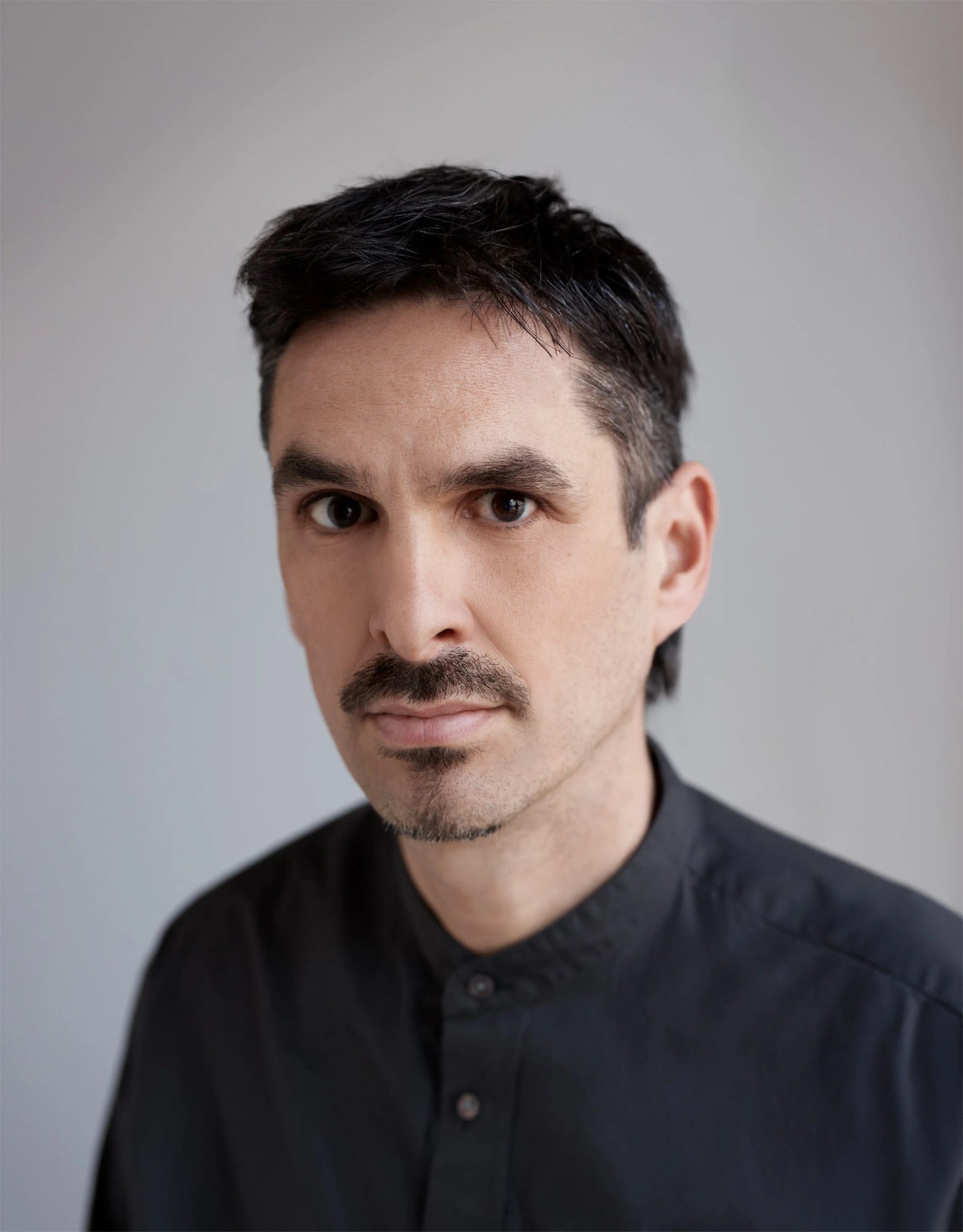 Close-up portrait of Raphael Ruz, a man with dark hair, a moustache and goatee, wearing a black button-up shirt and looking directly at the camera against a neutral background.
