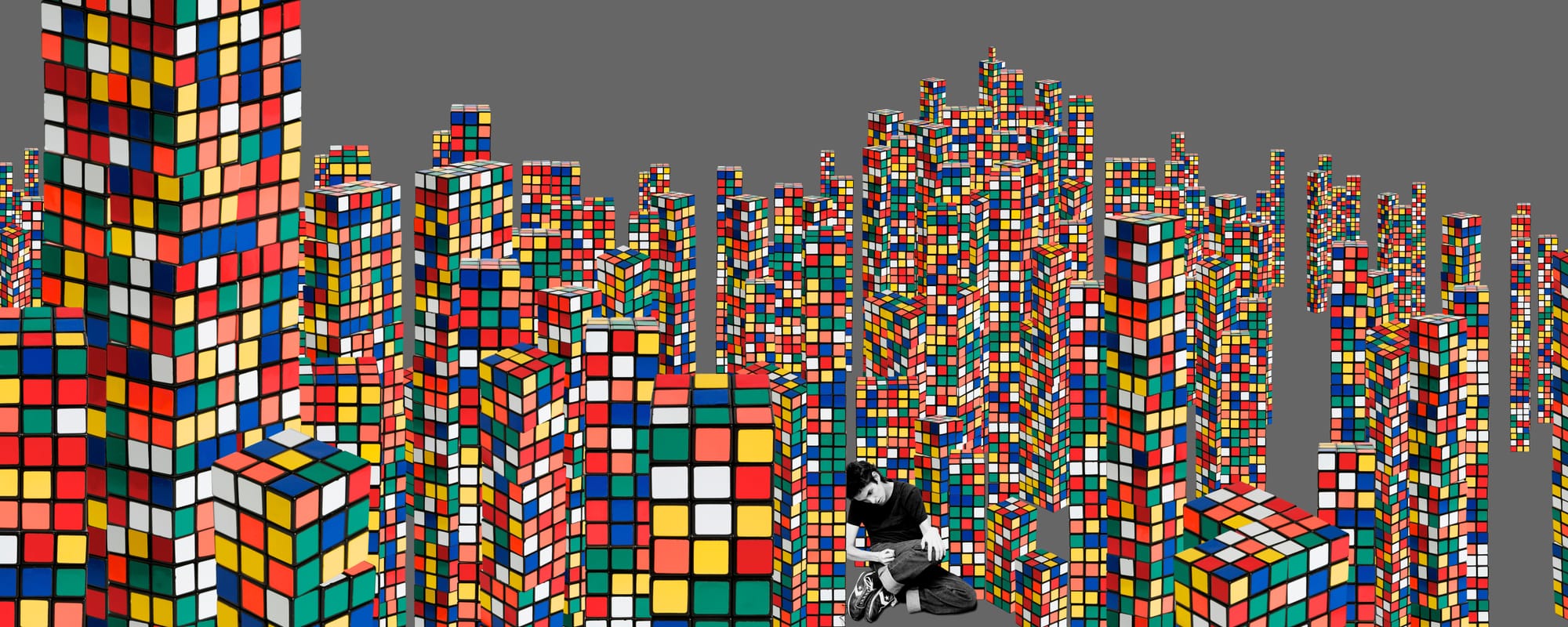A lone figure sitting amid a cityscape of giant, colourful Rubik’s cubes, against a grey background.