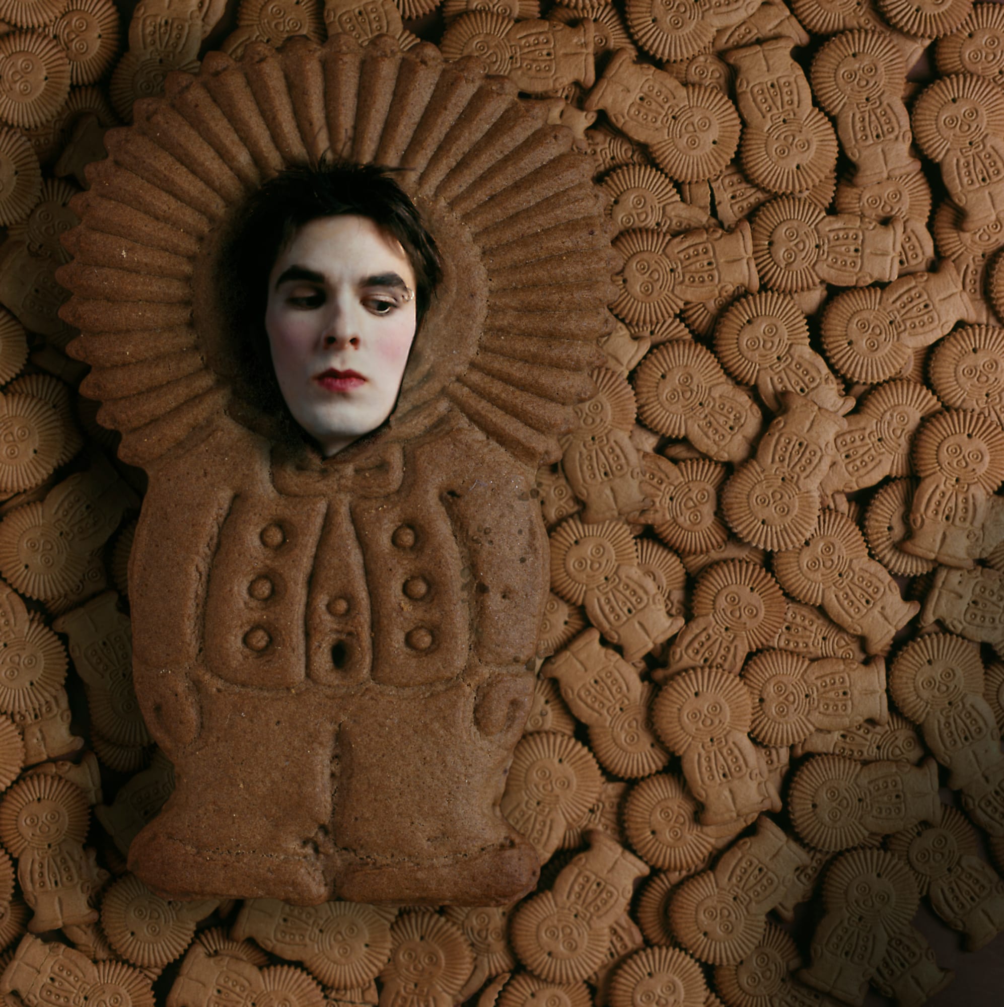Self-portrait as an infant in a brown ruffled dress with a high halo of ruffled fabric, before a rusted textured wall.
