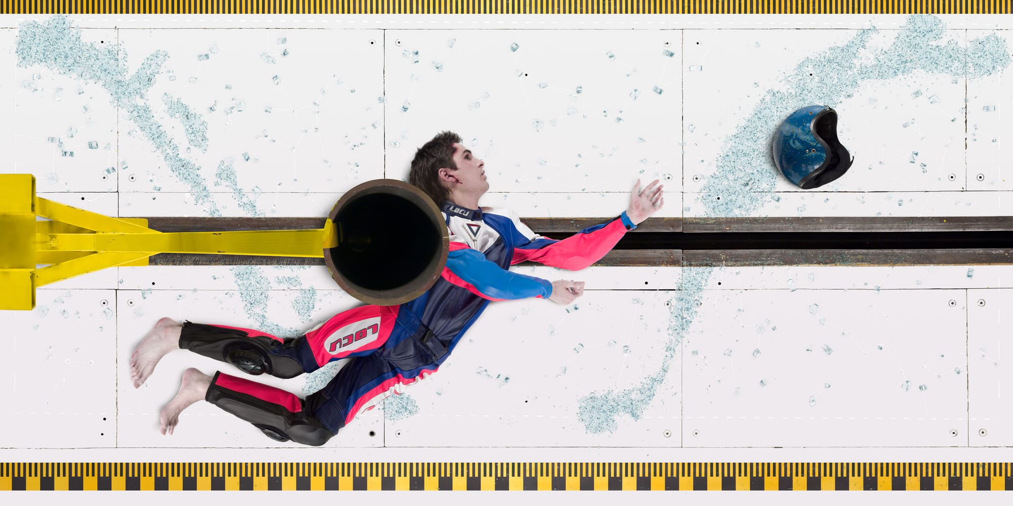 A figure in a colourful racing suit lying on the ground after a crash, a black helmet nearby, beside a yellow barrier and a track wall painted with a map of the earth.