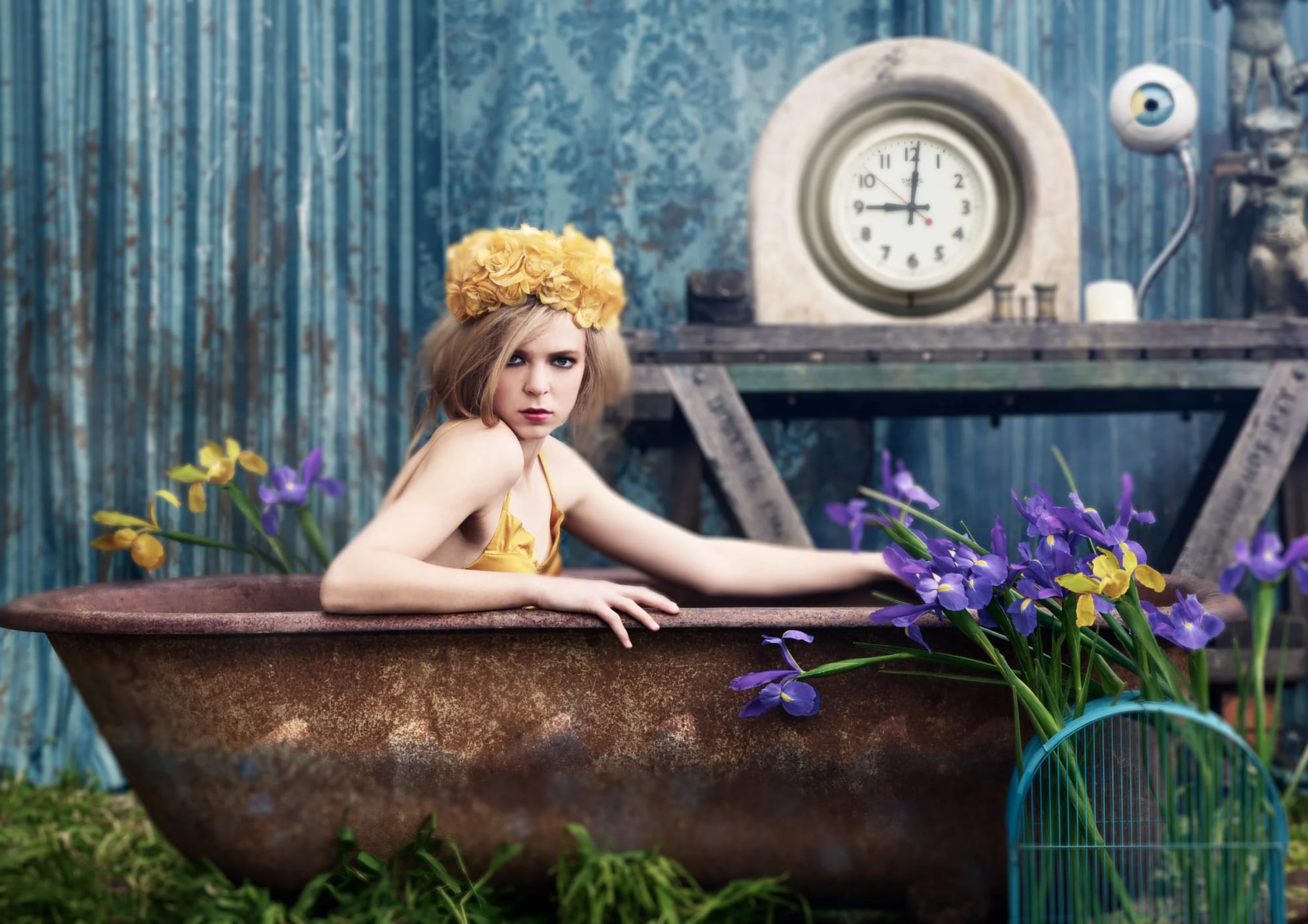 Fashion editorial: a woman with blonde hair, wearing a yellow dress and a yellow flower crown, sitting in a rusty bathtub with purple and yellow flowers surrounding her.