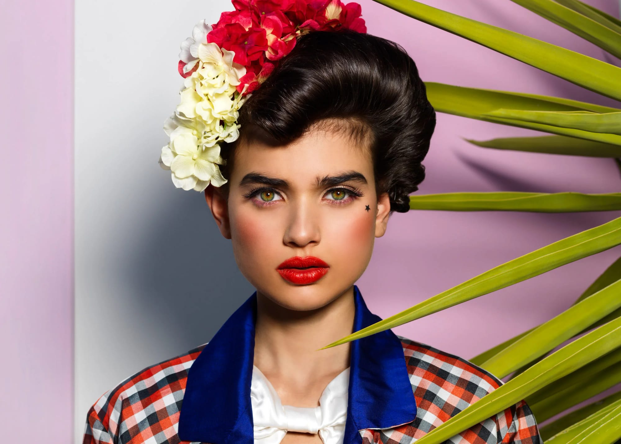 A woman with short, styled dark hair and green eyes, wearing bright red lipstick, a star tattoo near her right eye, a checkered shirt with a blue collar and white bow tie, and a large floral headpiece.