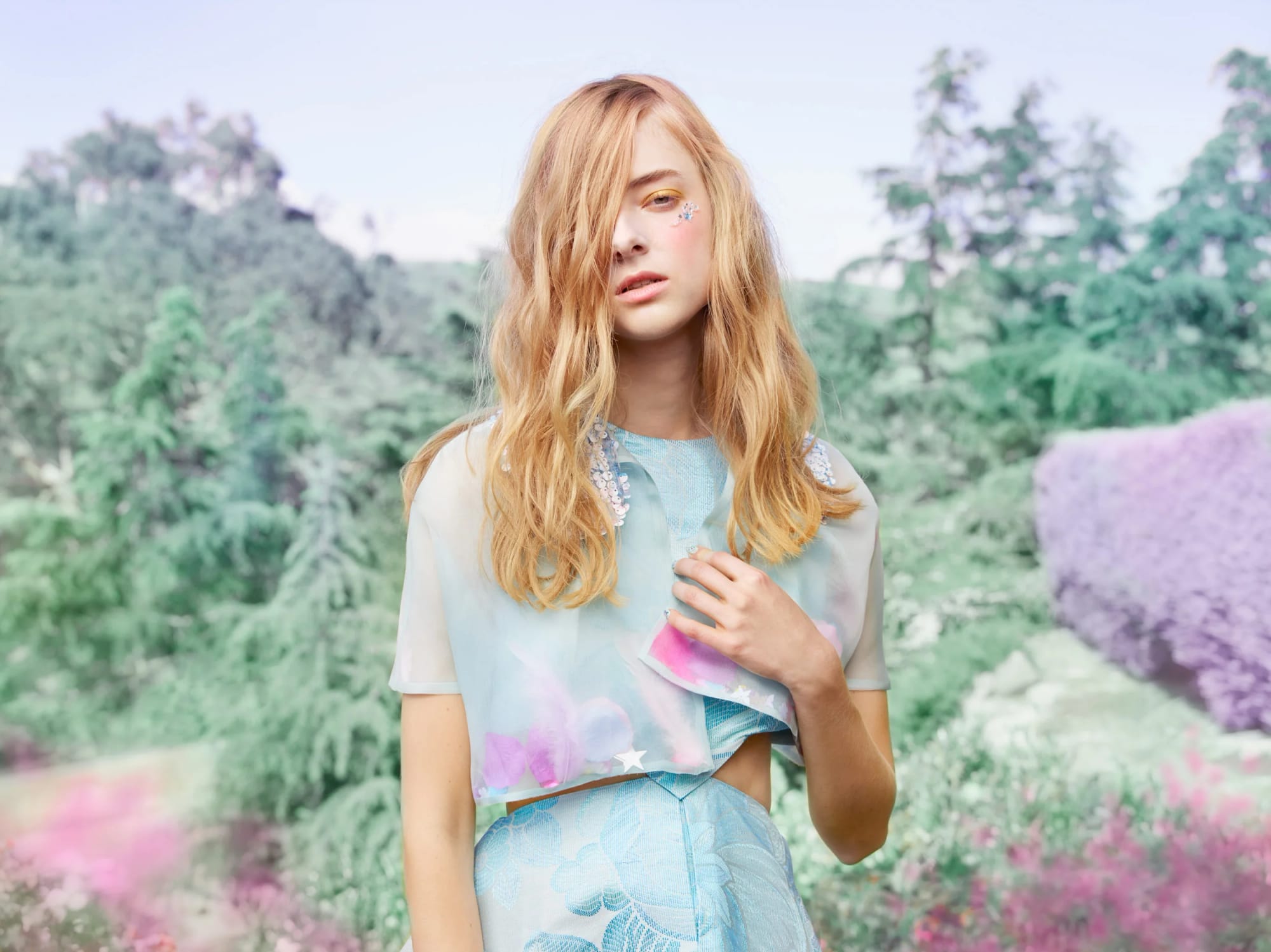 Young woman with long wavy red hair, wearing pastel-coloured clothing with floral patterns, standing in a garden with lush greenery and purple flowers.