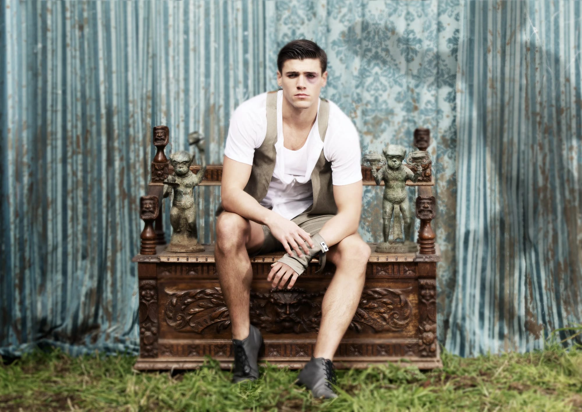 Young man sitting on a carved wooden throne with tribal statues, in front of blue patterned curtains and a grass floor.