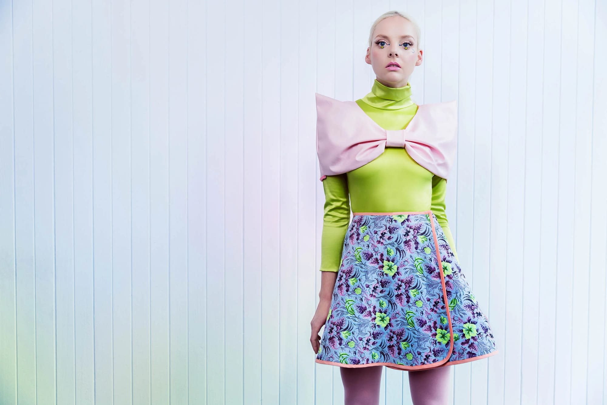 Young woman with platinum blonde hair and colourful makeup in a futuristic outfit with a large pink shoulder bow, lime green high-neck top, floral A-line skirt and pink tights.