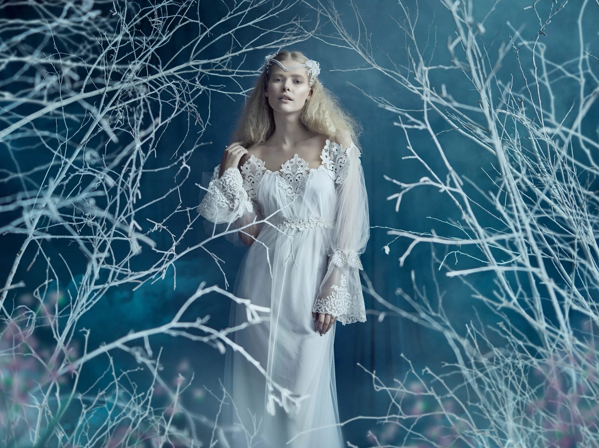 A woman dressed in a flowing white gown with lace details, standing among leafless white branches against a blue background.