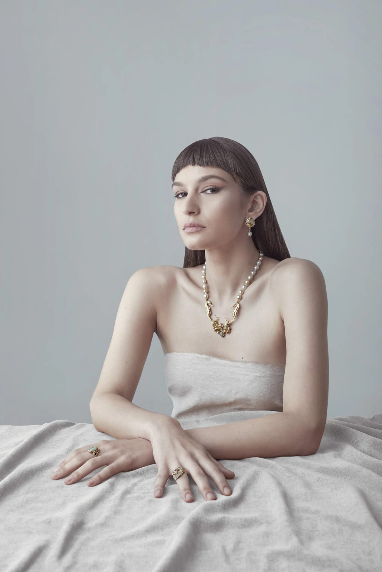 A woman with long dark hair and a blunt fringe wearing gold and pearl jewellery, sitting with her arm resting on a light surface, against a plain light grey background.