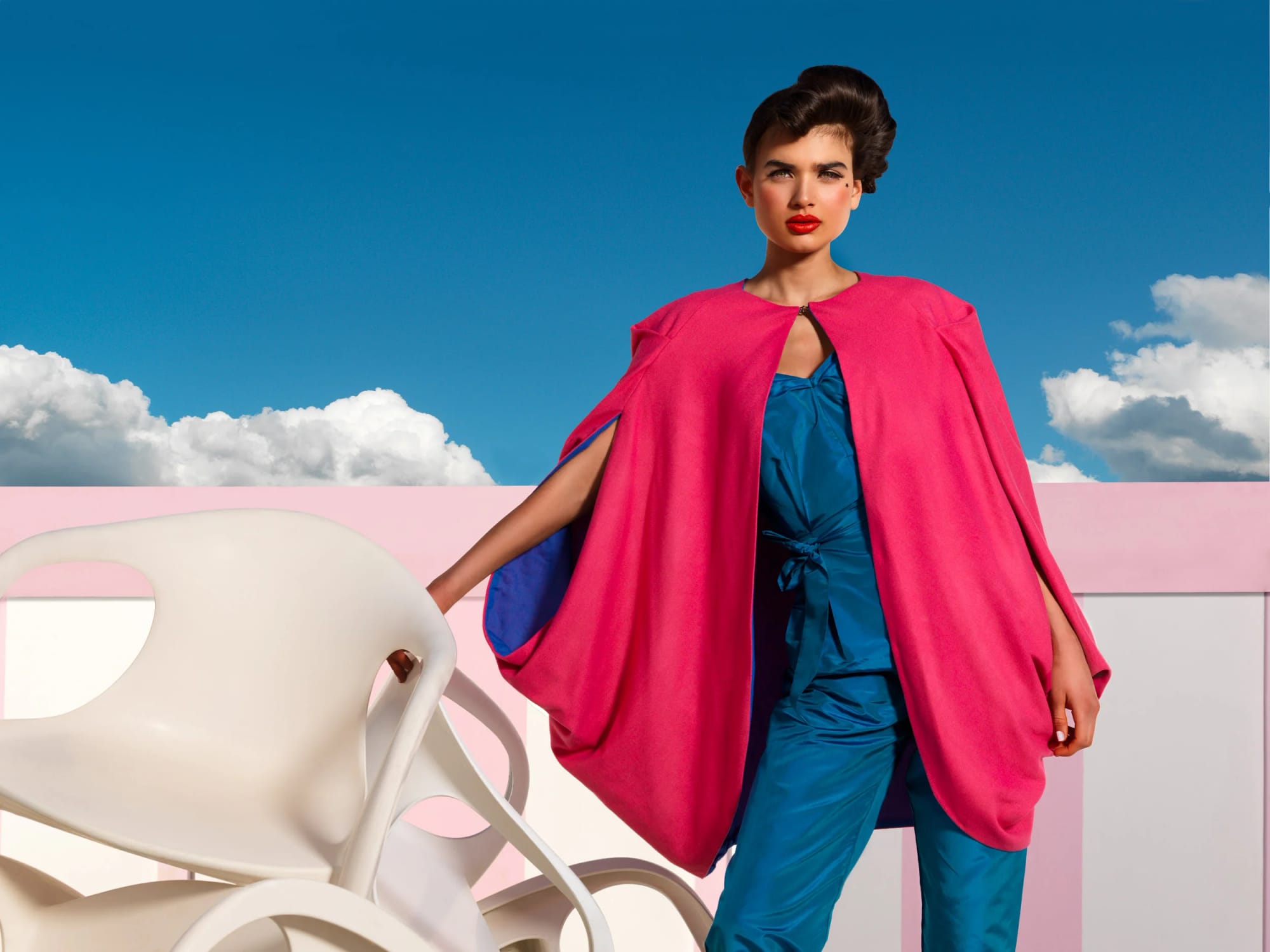 A woman with dark short hair and red lipstick standing outdoors against a blue sky with white clouds, wearing a bright pink cape and blue outfit.
