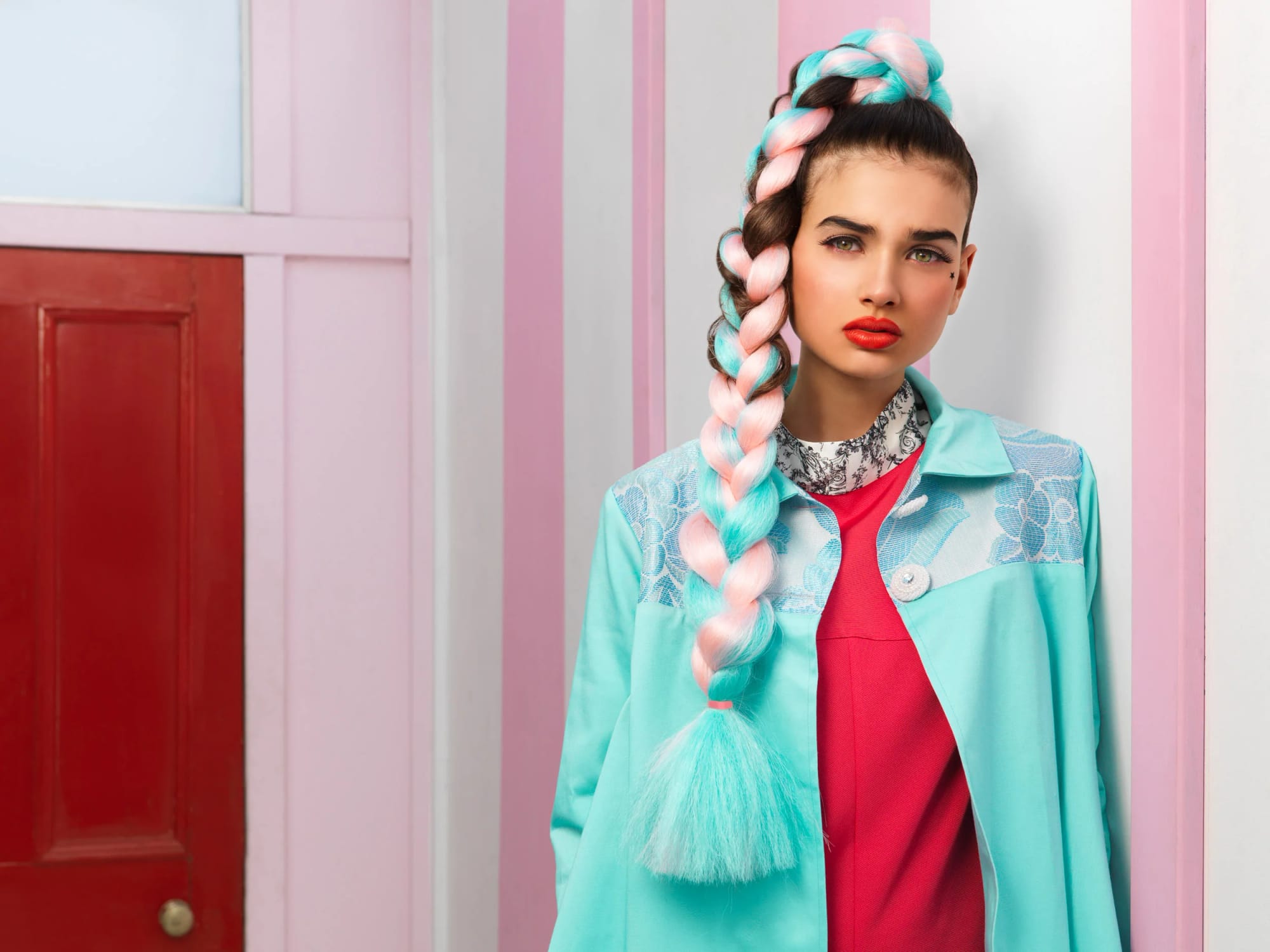 A young woman with colourful braided hair in shades of pink, blue and brown, wearing a teal jacket and red top, against pastel pink and white striped walls.