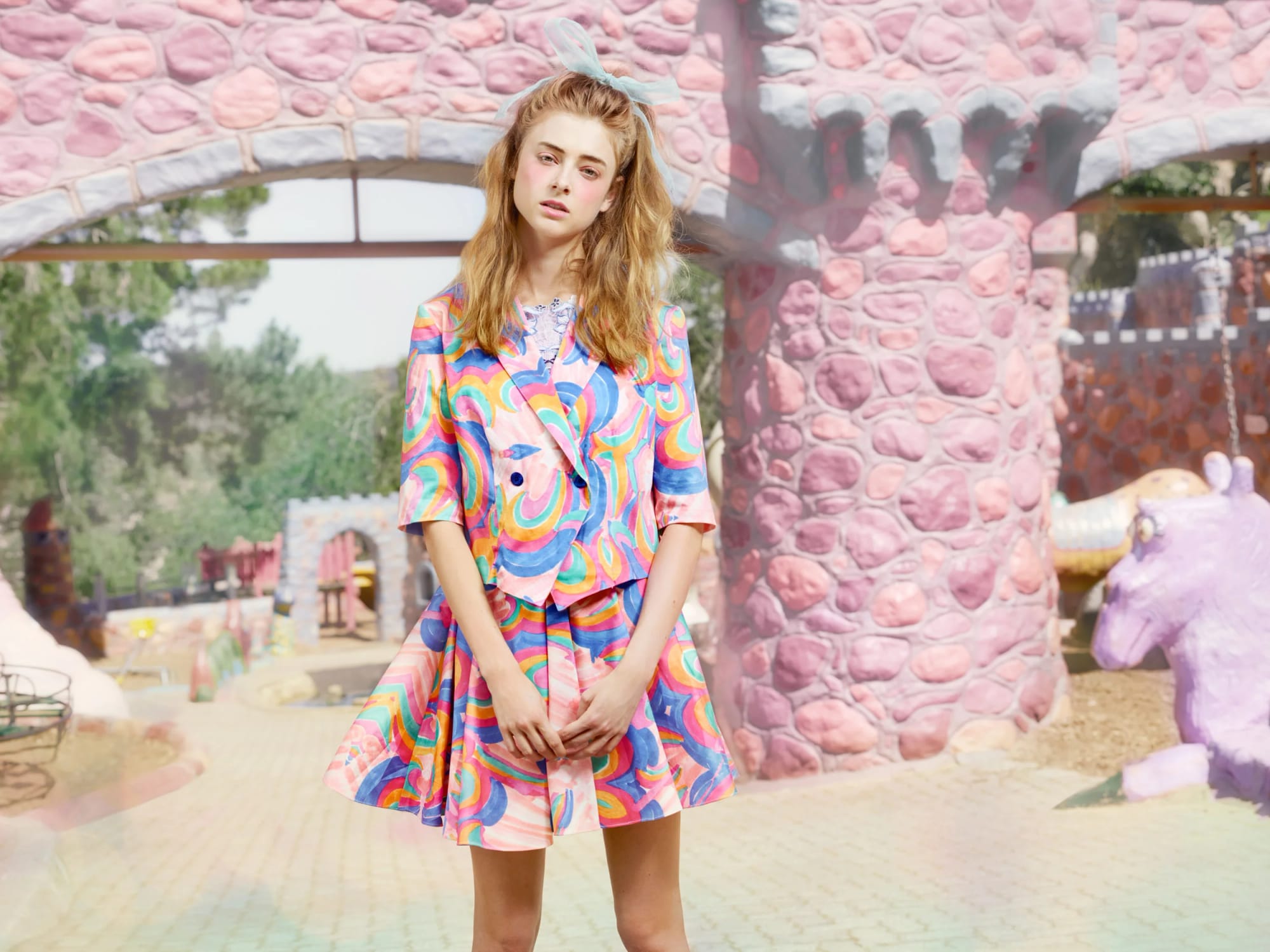 A young woman in a brightly coloured swirling-patterned outfit in front of a pink whimsical playground with castle-like structures and unicorn sculptures.