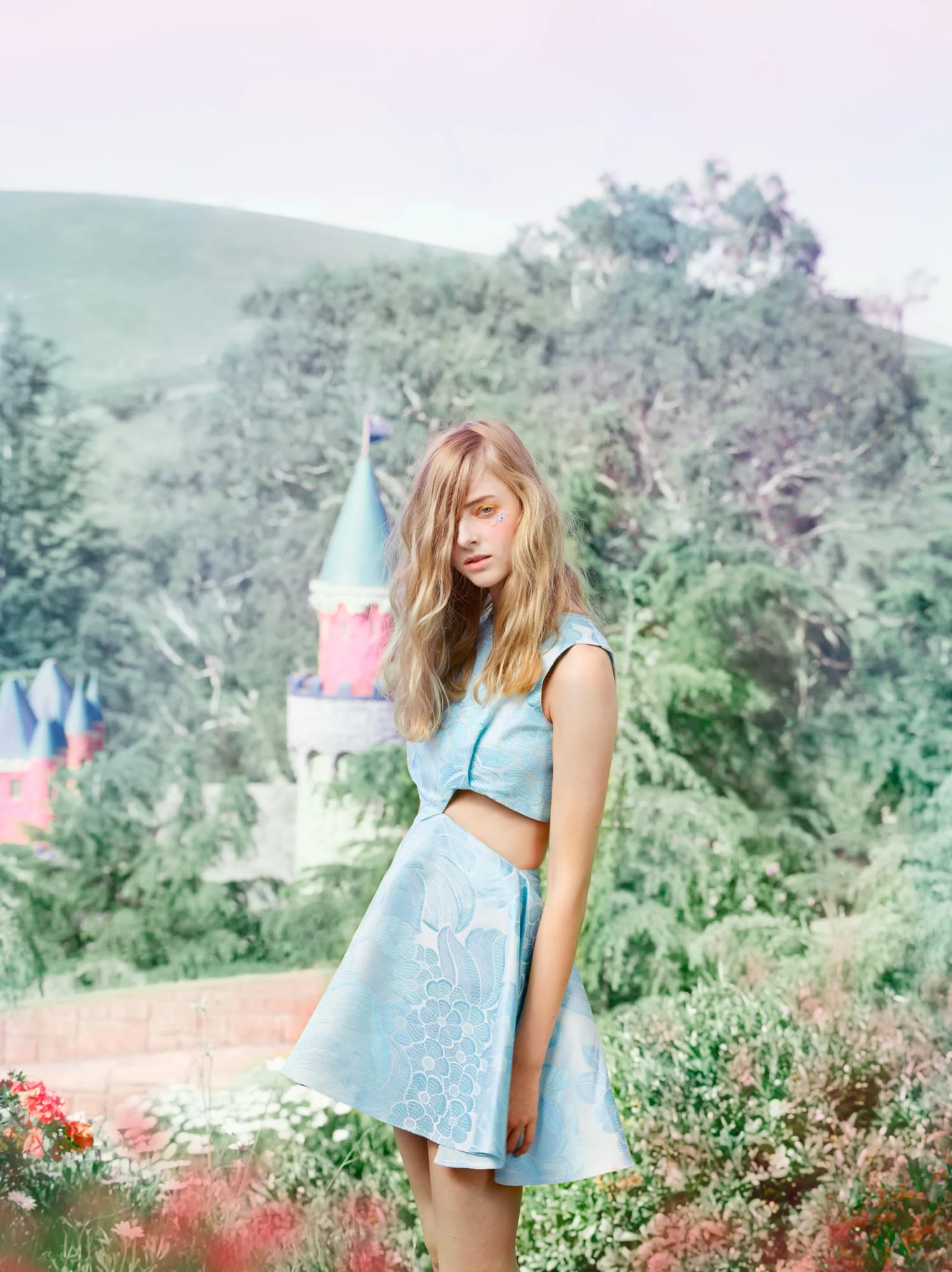A young woman with wavy blonde hair in front of a snowy mountain landscape with a castle, wearing a light blue patterned sleeveless dress with side cutouts.