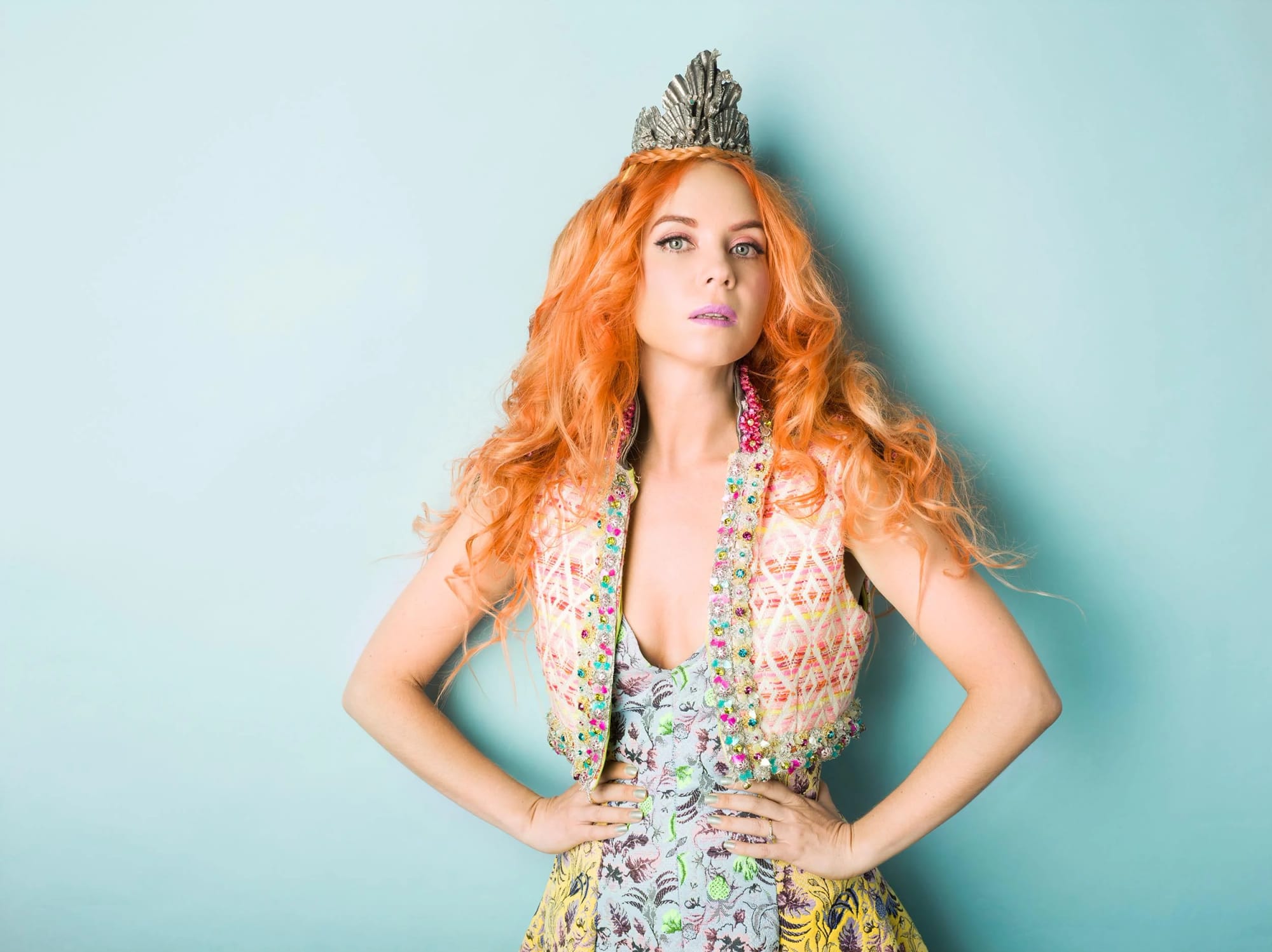 Portrait of Lady Petrova with curly red hair wearing a silver crown and a colourful patterned sleeveless dress, against a light blue background.