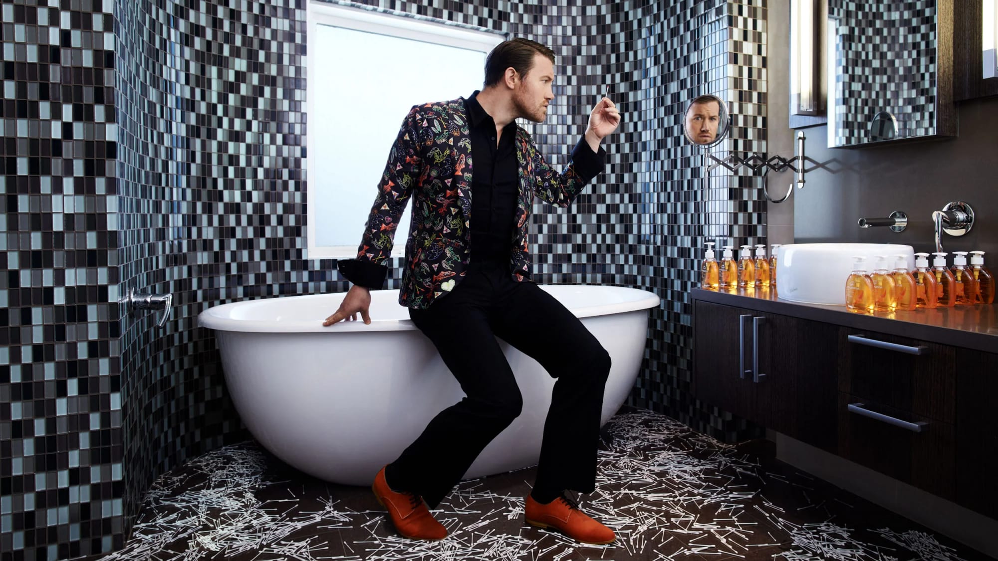 Portrait of Eddie Perfect sitting on a bathtub in a bathroom with black and white mosaic tiles, looking at his reflection in a small round mirror.