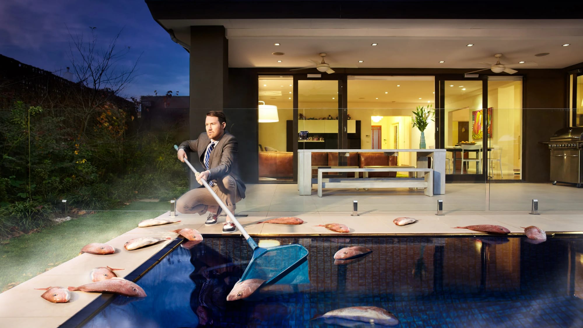 Portrait of Eddie Perfect in a suit cleaning a swimming pool with a net, surrounded by fish, in a backyard at night with a lit house behind.