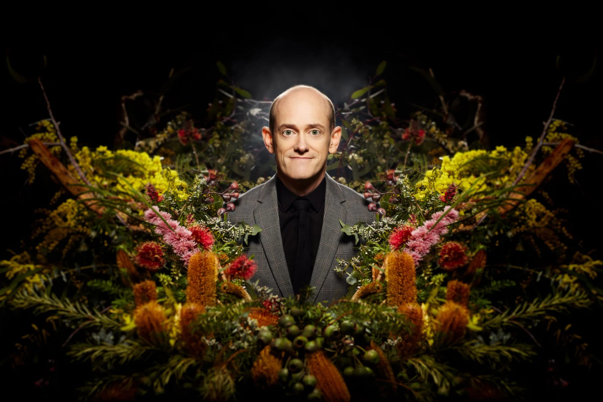A man in a suit and black shirt standing behind a large arrangement of colourful flowers against a dark background.