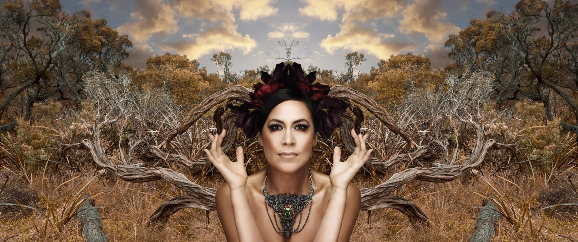 Kate Ceberano with dark hair, elaborate dark jewellery and a headpiece of dark purple flowers, posing before a mirror-like reflection in a forest scene.