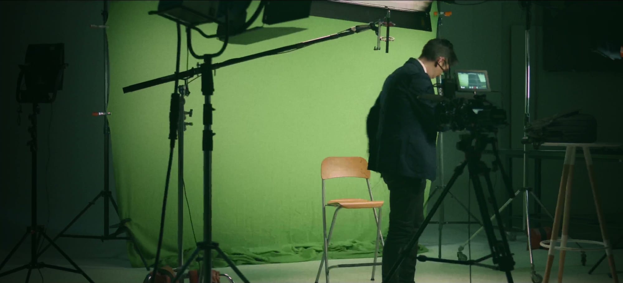 A green screen rigged in the corner of an office, two light stands and a wooden chair in front of it, and one man in a suit setting the camera up himself