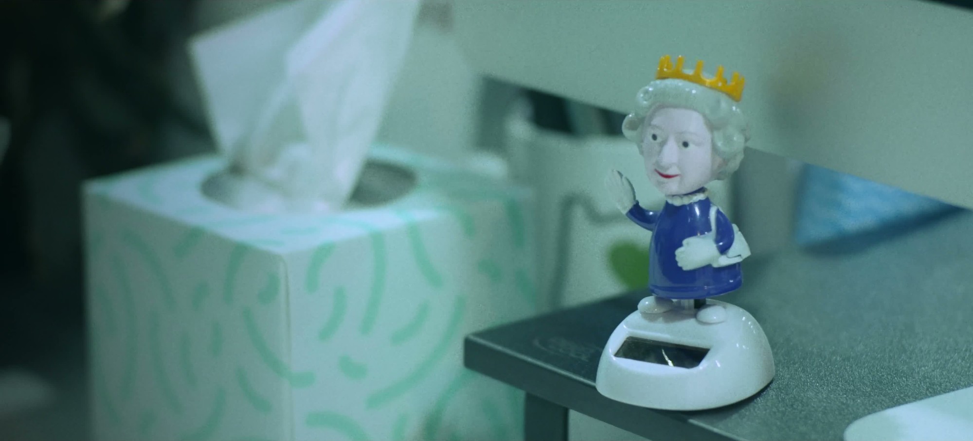 A small solar-powered Queen figurine waves beside a tissue box and a computer monitor on an office desk.