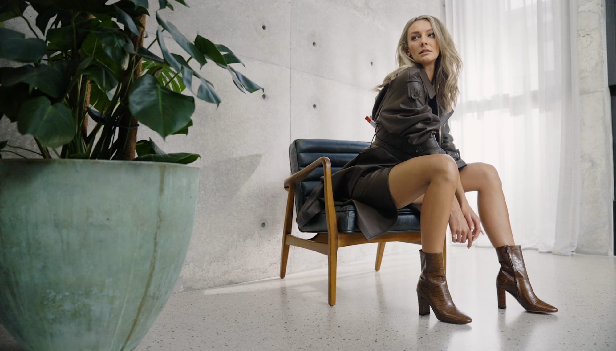 A model in a leather jacket and tan heeled boots, sitting back in a low armchair beside a large potted plant, daylight through the curtains behind her