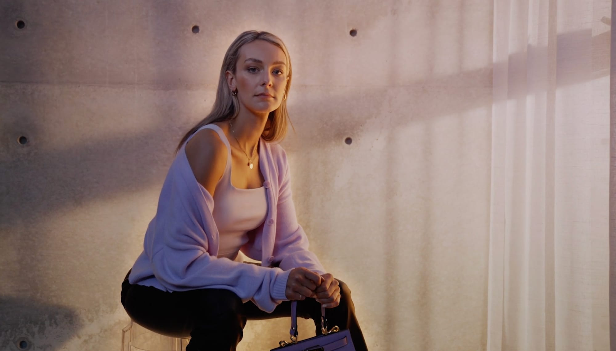 A model in a lilac cardigan crouched against a concrete wall, a purple handbag set down beside her feet