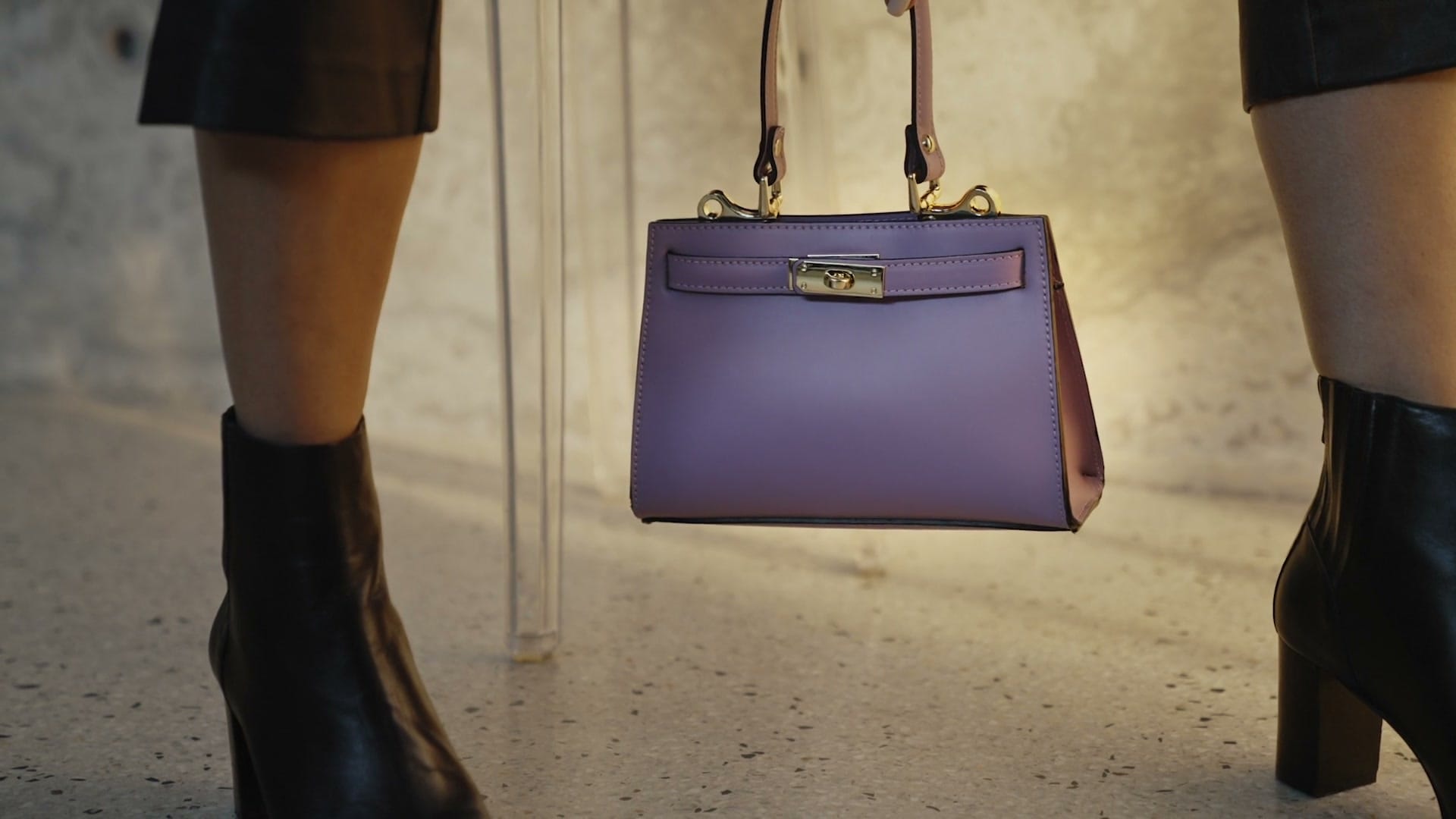 A lilac top-handle handbag held up at knee height between two models in black leather boots