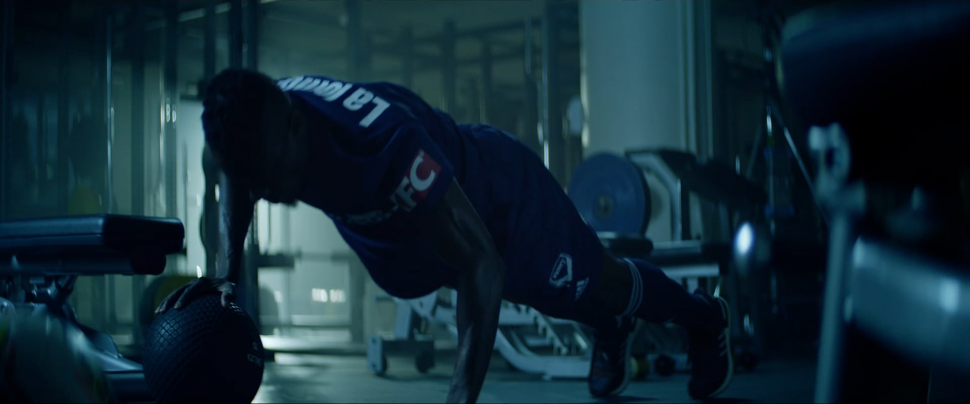 A Melbourne Victory player drives through a push-up over a medicine ball in a dark gym.