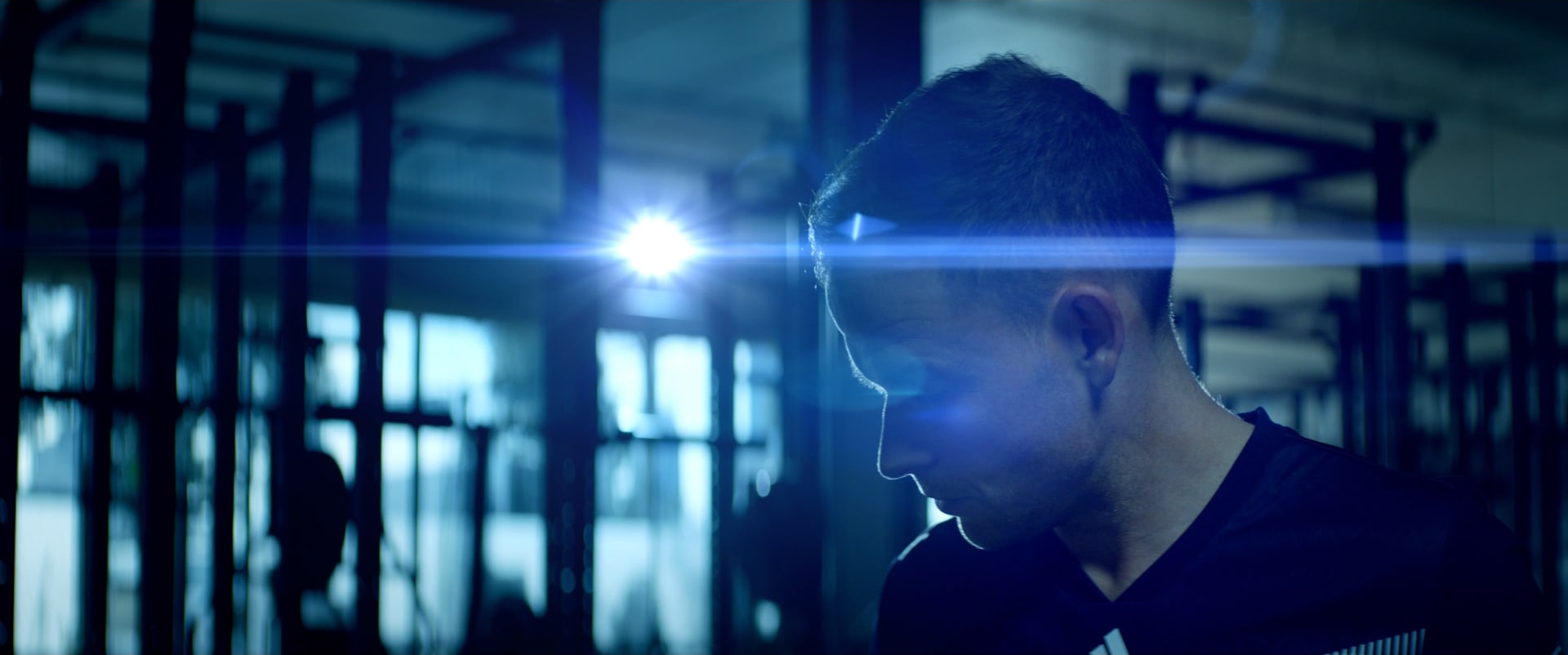 A player looks down, lit by a lens flare between the gym's steel racks.