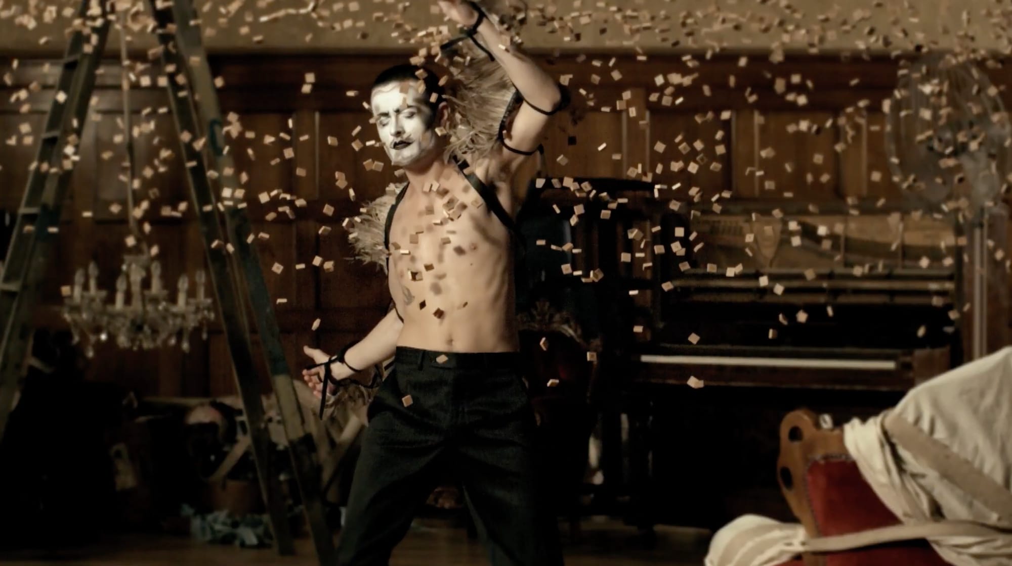 A shirtless performer in white face paint, one arm raised, as confetti falls through a wood-panelled hall