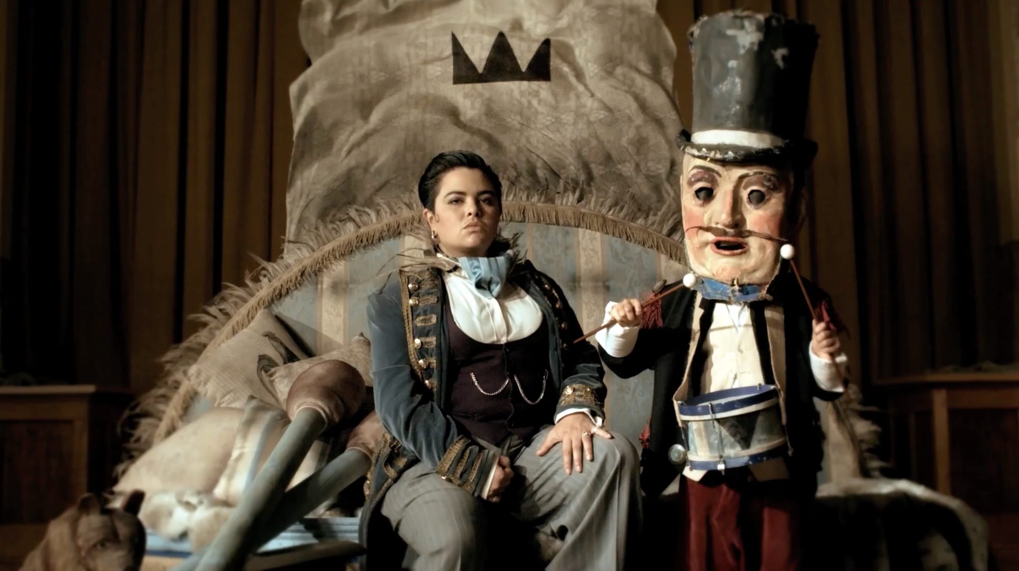 Two performers on a bed beneath a painted crown, one in a military jacket, the other in a top hat and a puppet mask