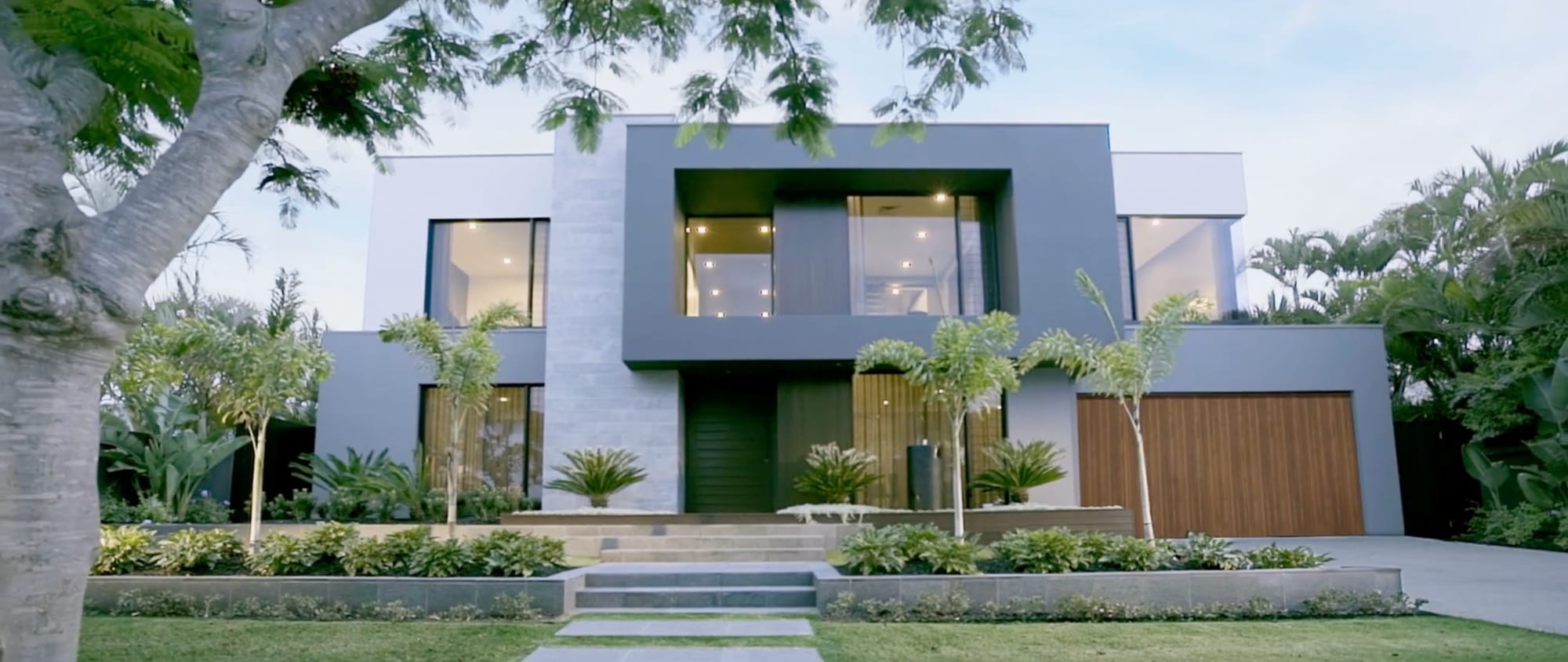 A two-storey house at dusk, lit from inside, palms and a clipped lawn across the front