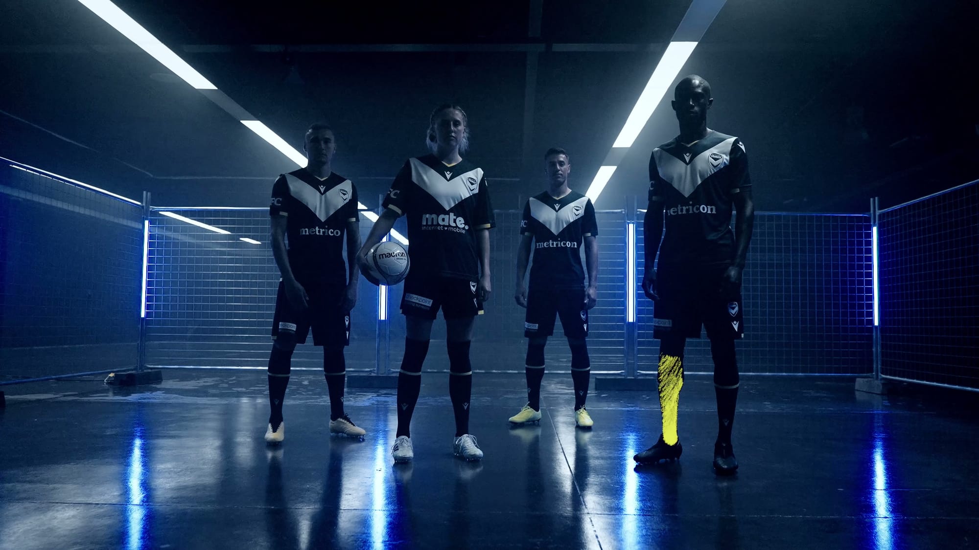 Four Melbourne Victory players standing across a tunnel set, one holding a ball, neon strips running down the walls behind them