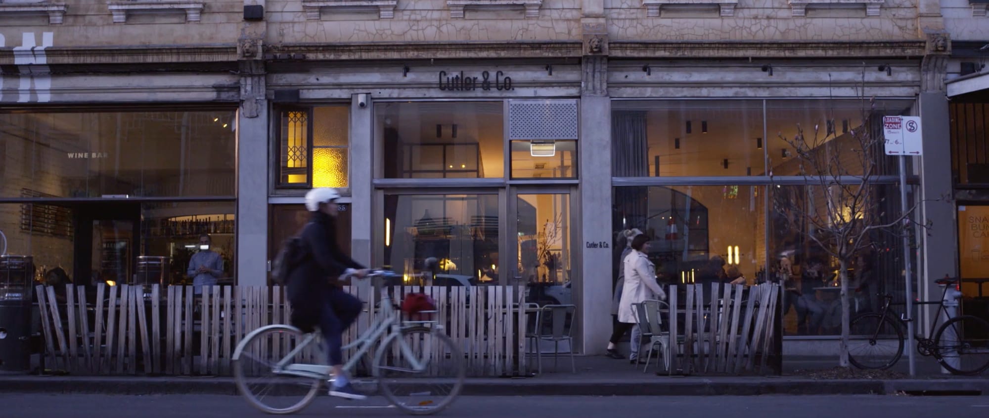 A shopfront on an East Melbourne street at dusk, warm light inside, a cyclist passing the window