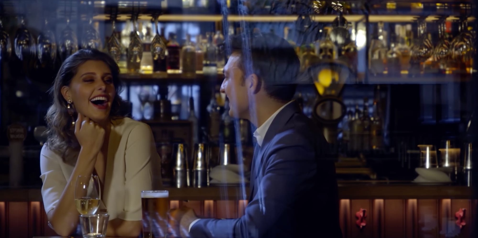 A woman laughing with a man in a suit across a bar, spirit bottles lit behind them