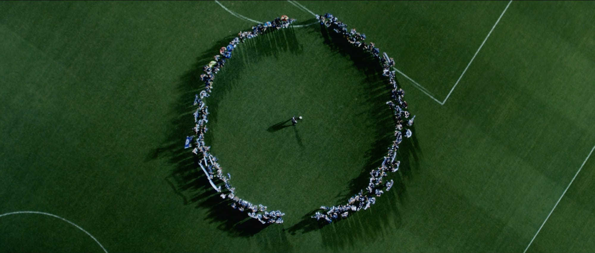 Shot from above, players and supporters standing shoulder to shoulder in a single ring on the pitch, forming a zero around one person at its centre