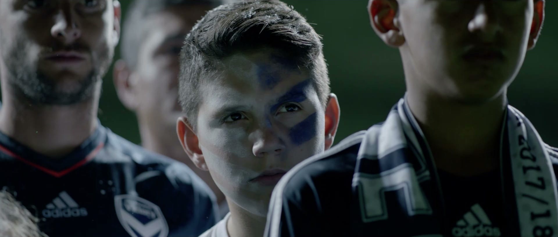 Three young supporters in Victory scarves and jerseys, blue face paint under their eyes, watching something off-camera