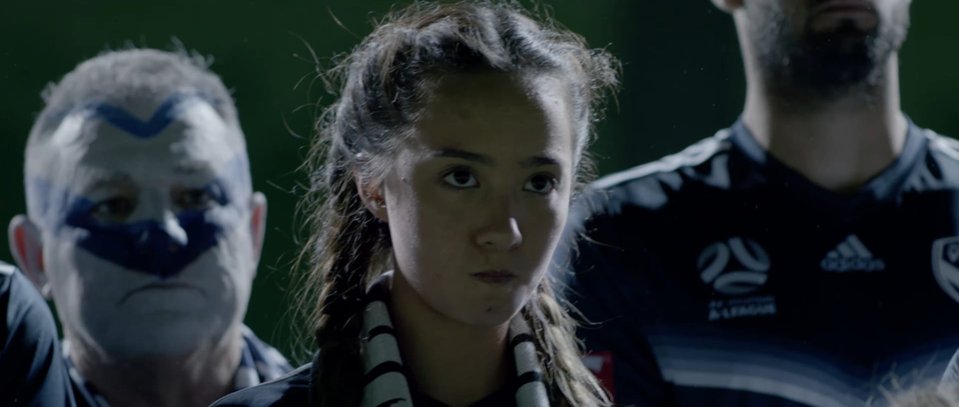 A young Melbourne Victory supporter with braided hair and a club scarf stands among fans with painted faces, looking ahead under stadium lights.