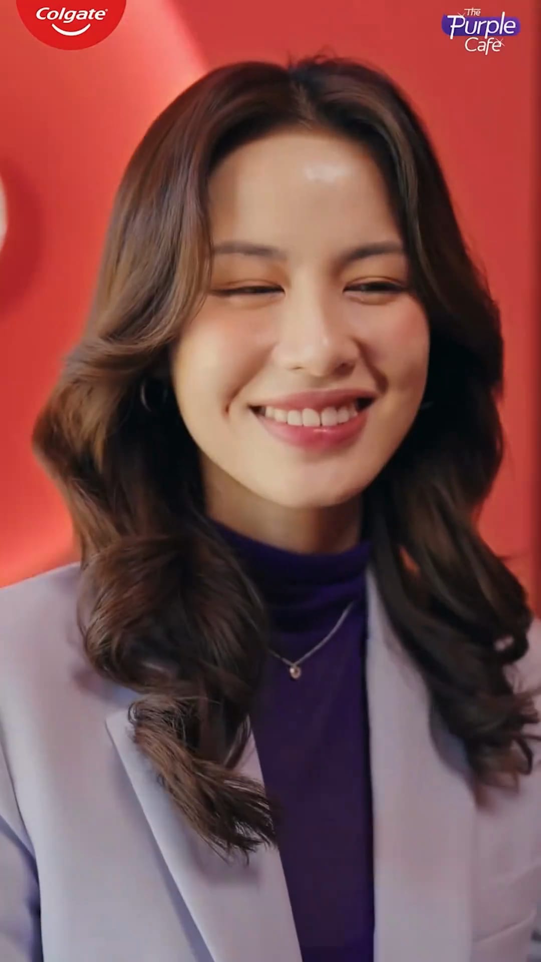 A lead from The Purple Café series smiles at the purple café table, in a lilac blazer, with the Colgate and Purple Café marks in frame.
